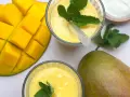Zwei Gläser cremiger Mango Lassi, garniert mit Minze, neben frischen Mangos und einem Schälchen Joghurt.