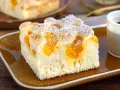 Ein Stück saftiger Buttermilchkuchen mit Mandarinen, bestäubt mit Puderzucker, auf einem braunen Teller neben einer Tasse Kaffee.