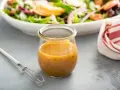 Ein Glas mit honig senf dressing steht neben einem kleinen Schneebesen. Im Hintergrund eine Schüssel mit Salat.