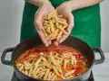 Hände geben Penne in einen Topf mit köchelnder Sauce für ein einfaches One-Pot-Chicken-Pasta-Gericht.