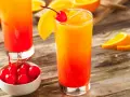 Ein erfrischender Tequila Sunrise mit Orangenscheibe und Kirsche, daneben eine Schale mit Kirschen.
