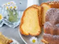 Saftiger Omas Joghurt Kuchen, frisch gebacken und mit Puderzucker bestäubt, daneben eine Zitronenscheibe und Gänseblümchen.