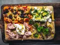 Vier verschiedene Beläge auf einer rechteckigen pizza al taglio: Oliven, Ricotta mit Kräutern, Mortadella mit Burrata und Kartoffeln mit Pilzen.