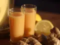 Zwei Gläser mit einem leuchtend orangen Ingwer Shot stehen neben frischem Ingwer und Zitronenscheiben.
