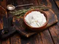 Cremiger, selbstgemachter Haferjoghurt in einer Holzschale, bereit zum Genießen. Ein Löffel und Kräuter liegen daneben.