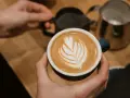 Cappuccino selber machen: Hände halten eine Tasse mit Latte Art. Im Hintergrund Milchkännchen und Sieb.