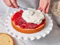 Eine Hand verteilt Sahne auf einer Marmeladenschicht eines Victoria Sponge Cake.
