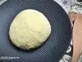 Ein runder Pizzateig auf schwarzer Platte wartet darauf, zu wissen, wie lange muss ein pizzateig ruhen, bevor er perfekt wird.