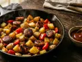 Heiß dampfende Bratwurst Kartoffel Pfanne mit Paprika, serviert in einer Gusseisenpfanne mit einer kleinen Schale Sauce.
