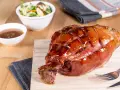 Glänzende, karamellisierte Schweinshaxe sous vide auf Holzbrett, daneben Schüssel mit Sauce und Salat.