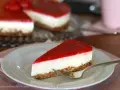 Ein Stück New York Cheesecake ohne Backen mit rotem Fruchttopping auf einem Teller, daneben ein ganzer Kuchen.
