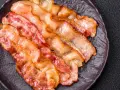 Knusprig gebratener Bacon im Ofen, auf einem dunklen Teller angerichtet.