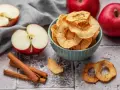 Leckere Apfelchips, die man ganz einfach selber machen kann, in einer Schüssel mit frischen Äpfeln und Zimtstangen.