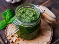 Frisches Basilikum Pesto in einem Glas, umgeben von Pinienkernen und Basilikumblättern.