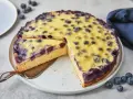 Ein saftiger Heidelbeerkuchen mit einer cremigen Vanillepudding-Schicht und frischen Heidelbeeren. Ein Stück wird gerade herausgeschnitten.