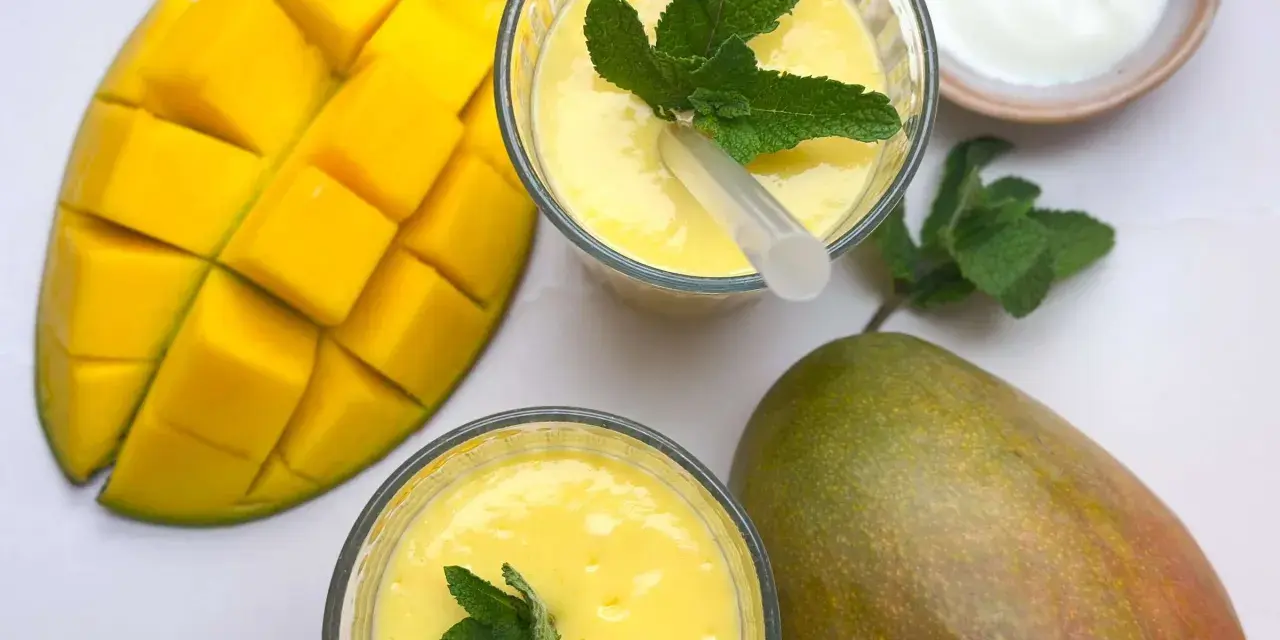 Zwei Gläser cremiger Mango Lassi, garniert mit Minze, neben frischen Mangos und einem Schälchen Joghurt.