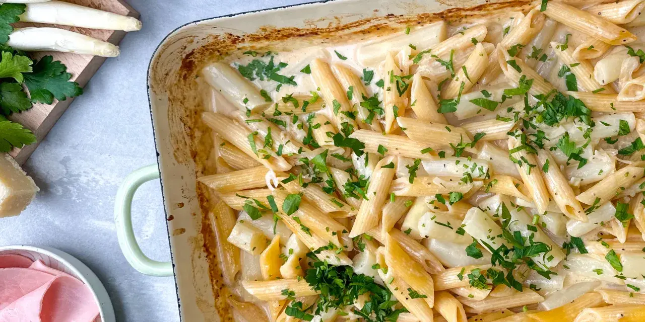Cremige Spargel Pasta mit frischer Petersilie in einer Auflaufform. Dazu liegen Spargelstangen, Schinken und Parmesan bereit.