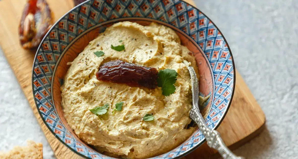 Cremiger Dattel Dip in einer bunten Schale, garniert mit einer Dattel und Petersilie. Dazu Brot und weitere Datteln auf einem Holzbrett.