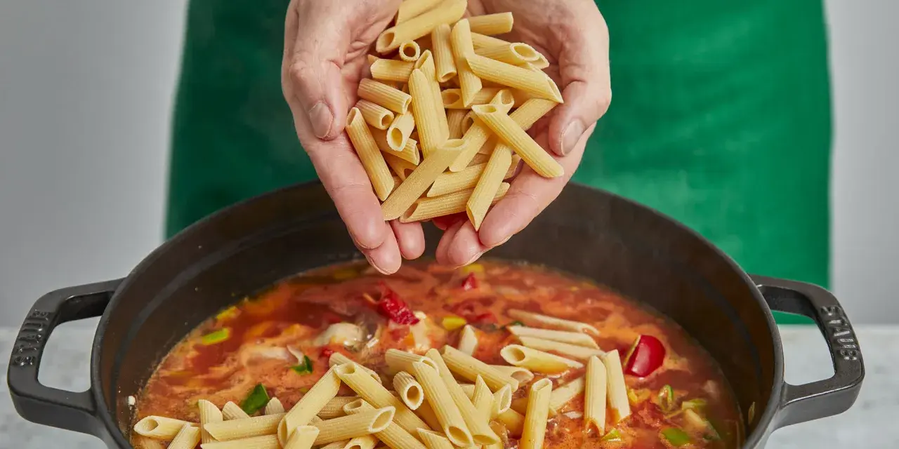 Hände geben Penne in einen Topf mit köchelnder Sauce für ein einfaches One-Pot-Chicken-Pasta-Gericht.