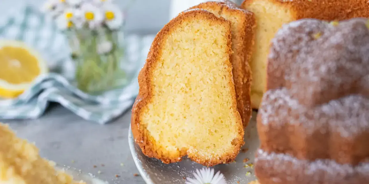 Saftiger Omas Joghurt Kuchen, frisch gebacken und mit Puderzucker bestäubt, daneben eine Zitronenscheibe und Gänseblümchen.