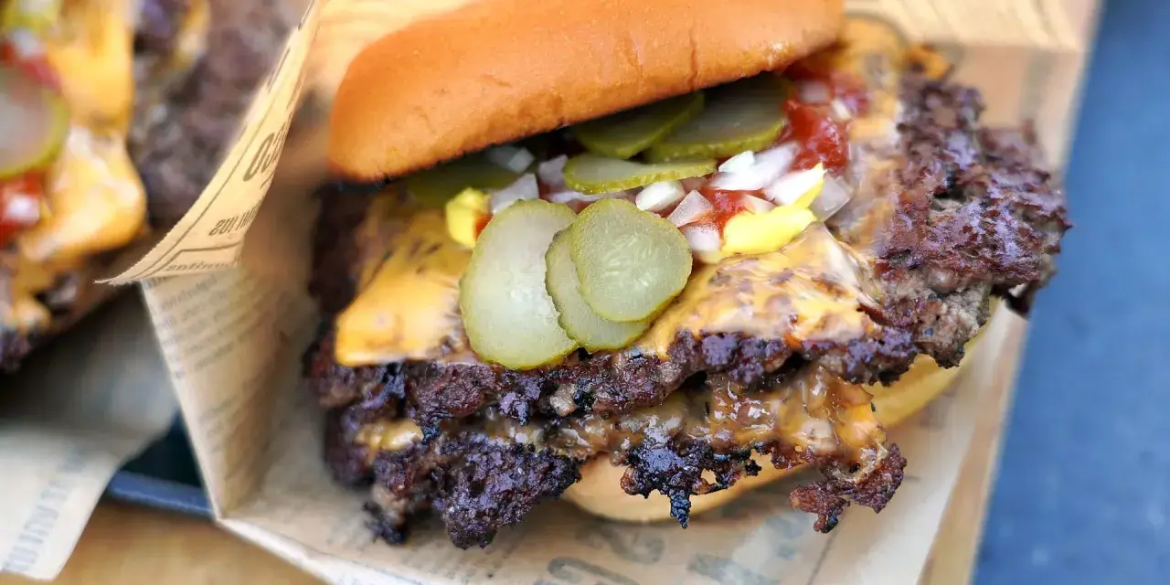 Saftiger Smash Burger mit geschmolzenem Käse, Gurken, Zwiebeln und Ketchup auf einem Brötchen, eingewickelt in Papier.