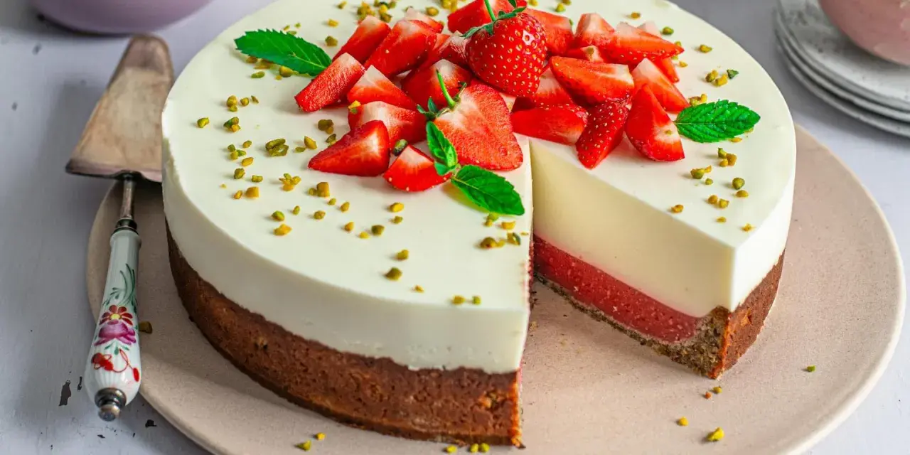 Erdbeer Joghurt Torte mit frischen Erdbeeren und Pistazien garniert. Ein Stück ist angeschnitten und zeigt die cremige Schicht.
