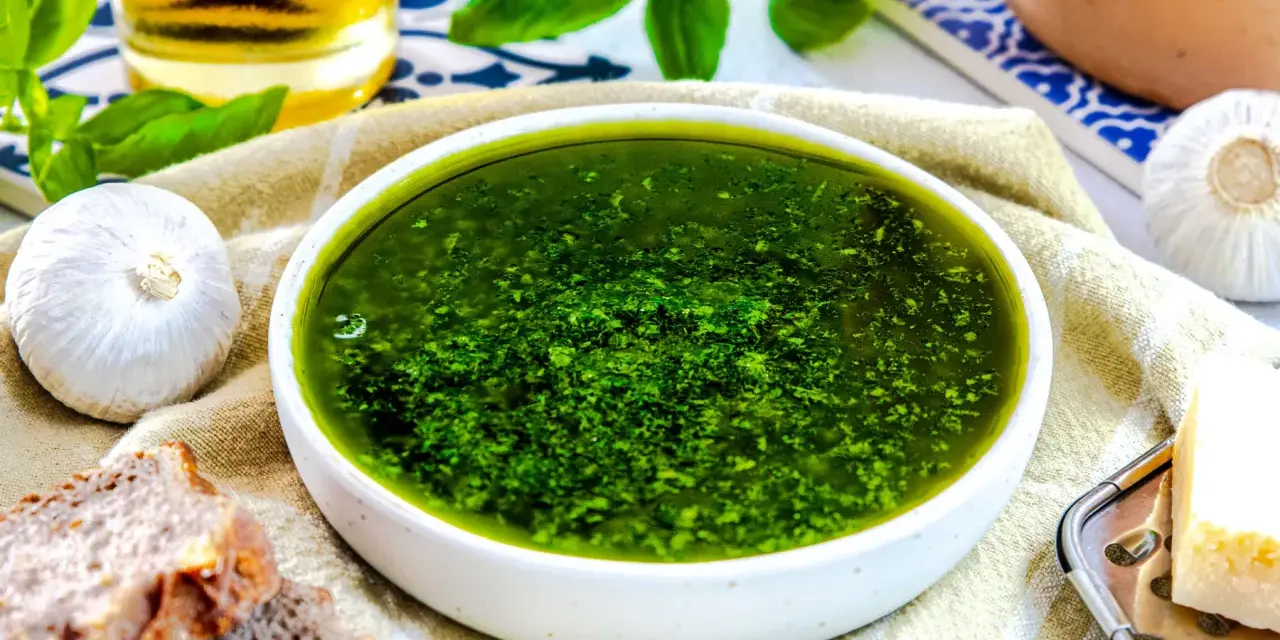 Frisches, grünes Pesto ohne Pinienkerne in einer Schüssel, daneben Knoblauch, Brot und Parmesan.