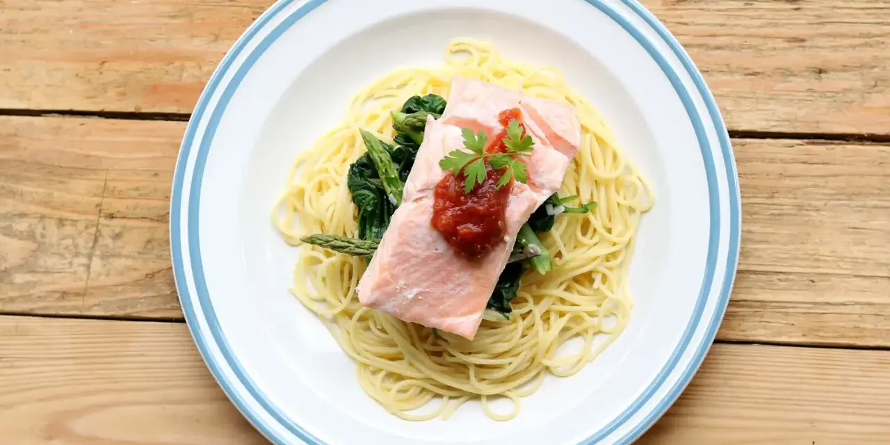 Ein Teller mit One Pot Lachs Pasta, garniert mit Petersilie und Tomatensauce, daneben Spargel und Spinat auf einem Holztisch.