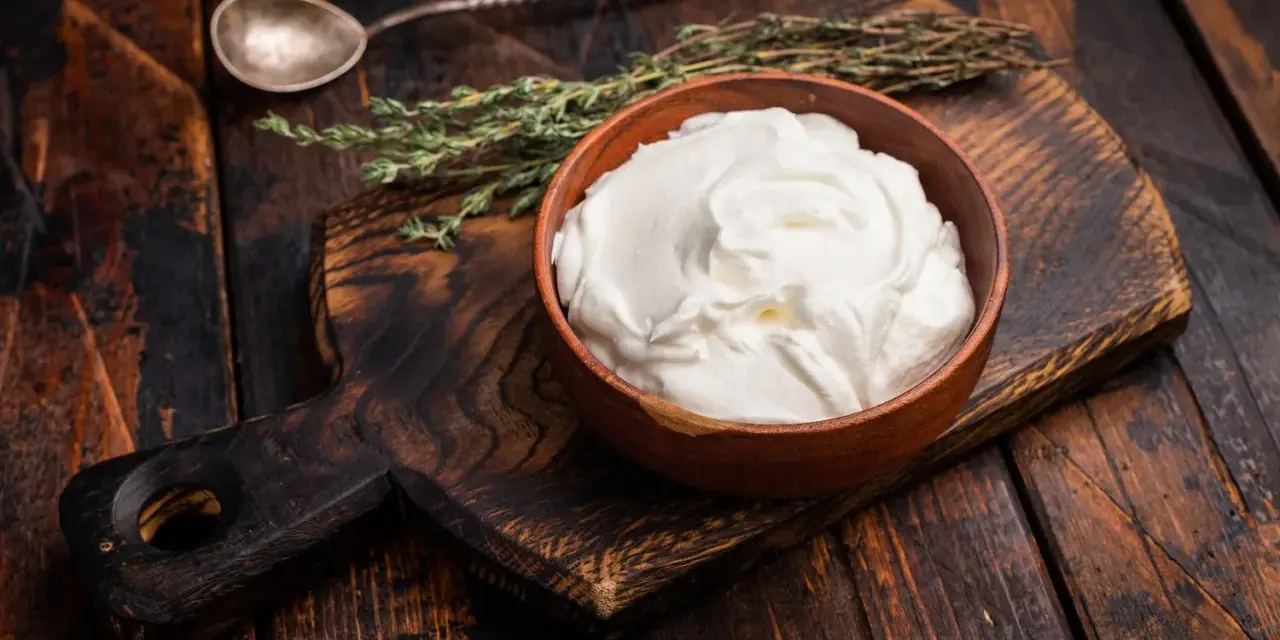 Cremiger, selbstgemachter Haferjoghurt in einer Holzschale, bereit zum Genießen. Ein Löffel und Kräuter liegen daneben.