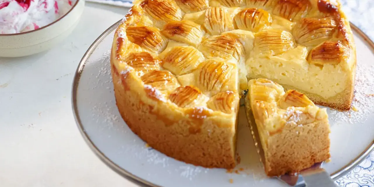 Ein köstlicher, schneller Apfelkuchen mit Schmand und Pudding, verziert mit Apfelscheiben und Puderzucker. Daneben eine Schüssel mit Beeren-Sahne.