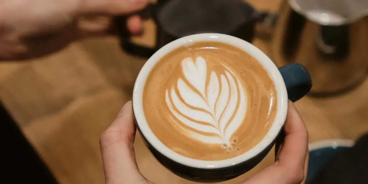 Cappuccino selber machen: Hände halten eine Tasse mit Latte Art. Im Hintergrund Milchkännchen und Sieb.