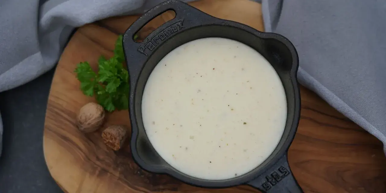 Cremige Béchamelsauce in einer kleinen Gusseisenpfanne, garniert mit Petersilie und Muskatnüssen.
