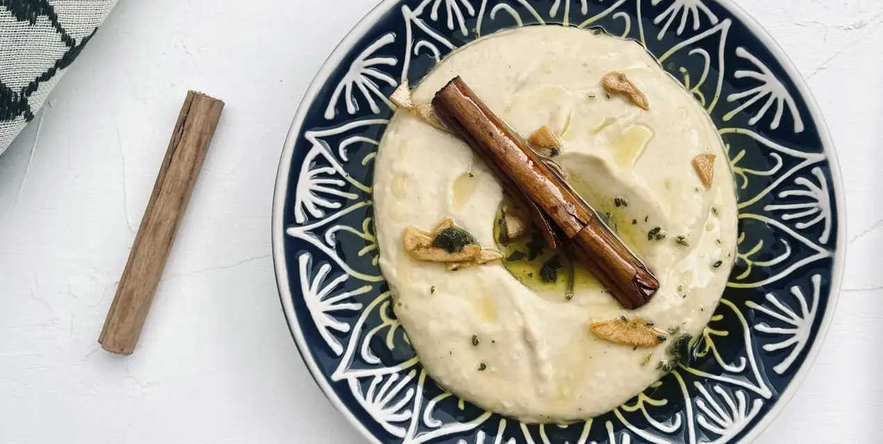 Cremiger Hummus, selbst gemacht, mit Zimtstange und Knoblauch garniert auf einem blau-weißen Teller.
