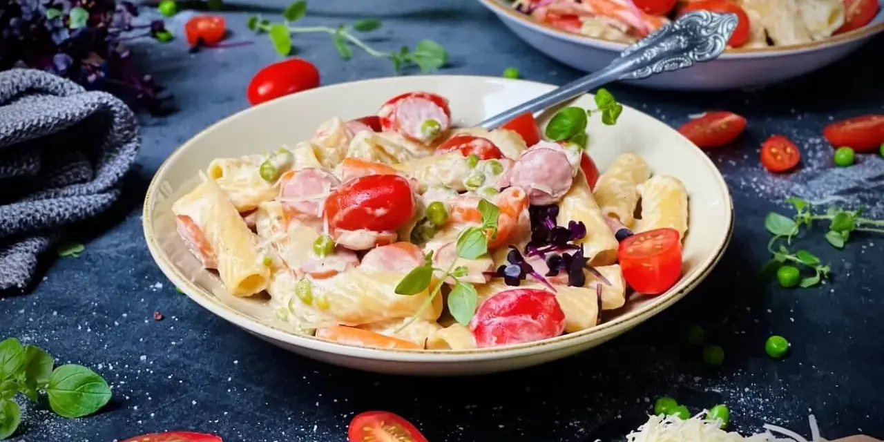 Cremige One Pot Pasta mit Würstchen, Erbsen und Kirschtomaten, garniert mit frischen Kräutern und geriebenem Käse.