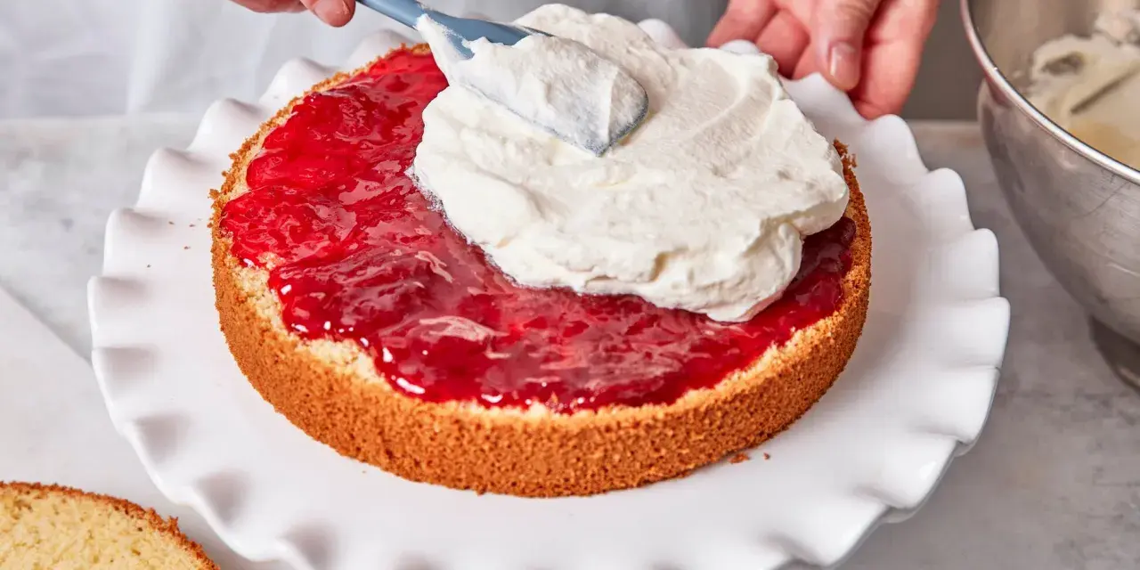 Eine Hand verteilt Sahne auf einer Marmeladenschicht eines Victoria Sponge Cake.
