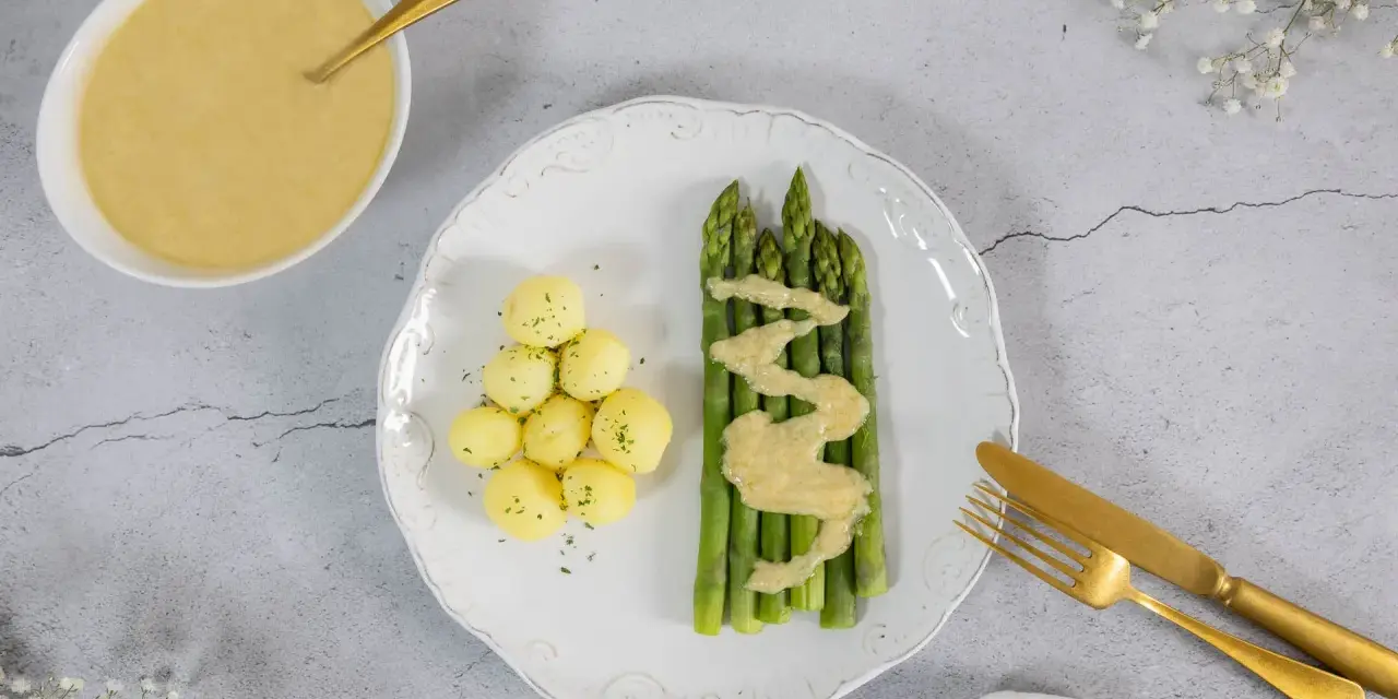 Spargel mit kleinen Kartoffeln und einer cremigen, veganen Sauce Hollandaise. Ein goldener Löffel liegt in der Sauce.