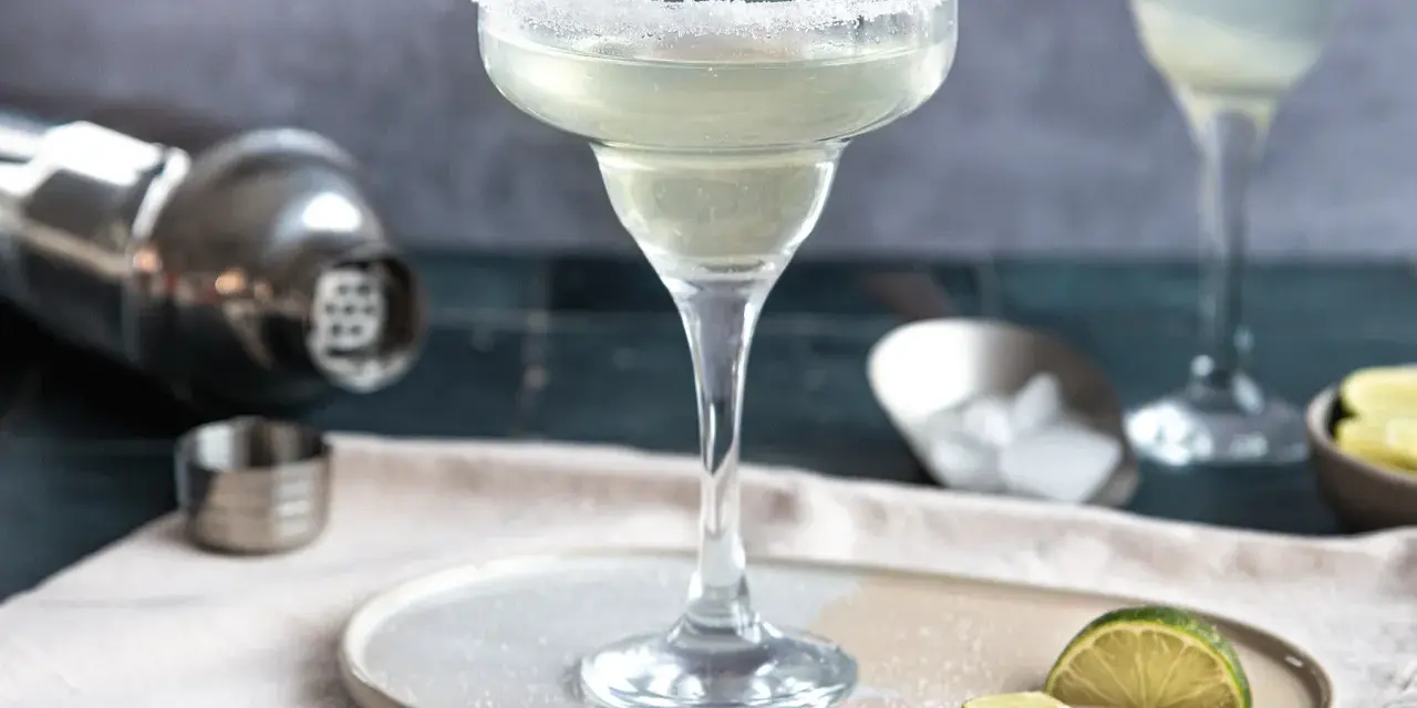 Ein perfekt zubereitetes Margarita-Rezept: Ein Glas mit Salzrand, daneben Limettenspalten, Eiswürfel und ein Shaker.