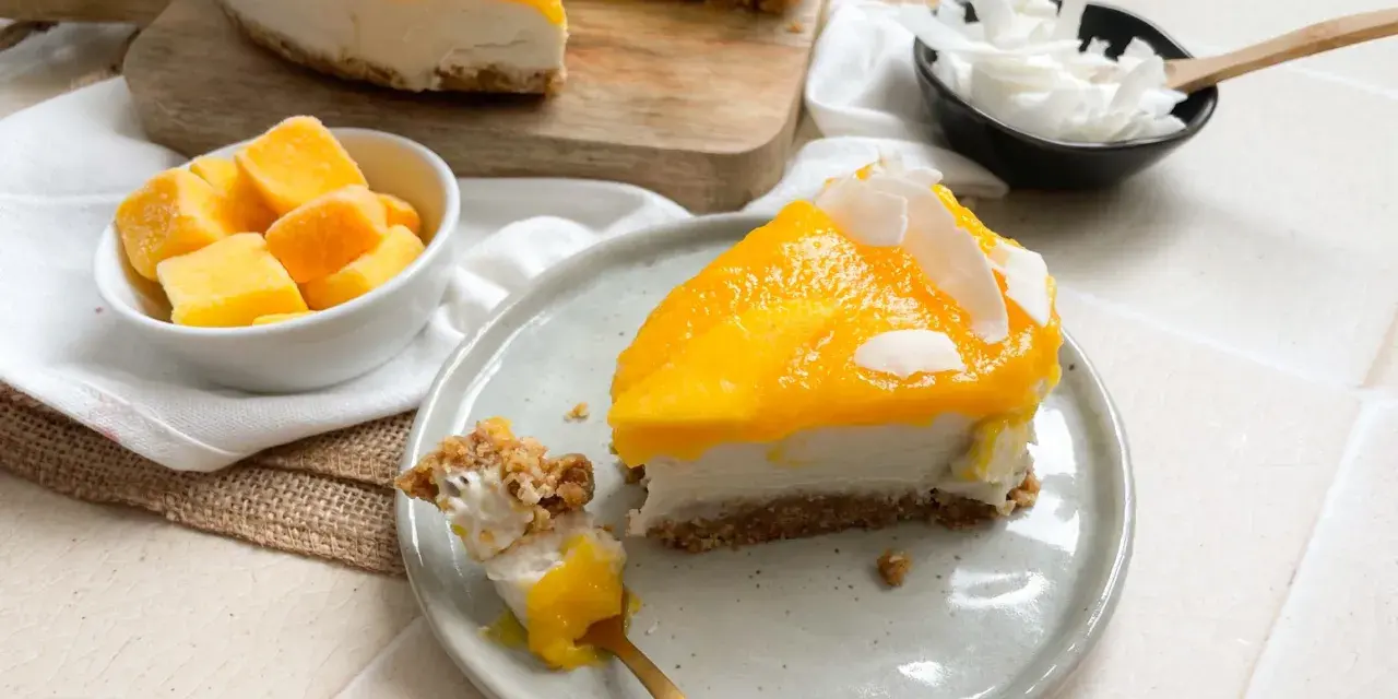 Ein cremiges Mango Cheesecake ohne Backen, garniert mit Kokosflocken. Ein Stück wird gerade mit einem Löffel probiert.