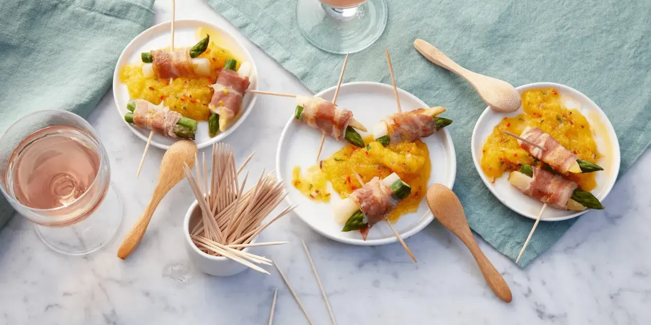 Leckere Spargel im Speckmantel auf gelbem Chutney, serviert auf kleinen Tellern mit Holzspießen und Roséwein.