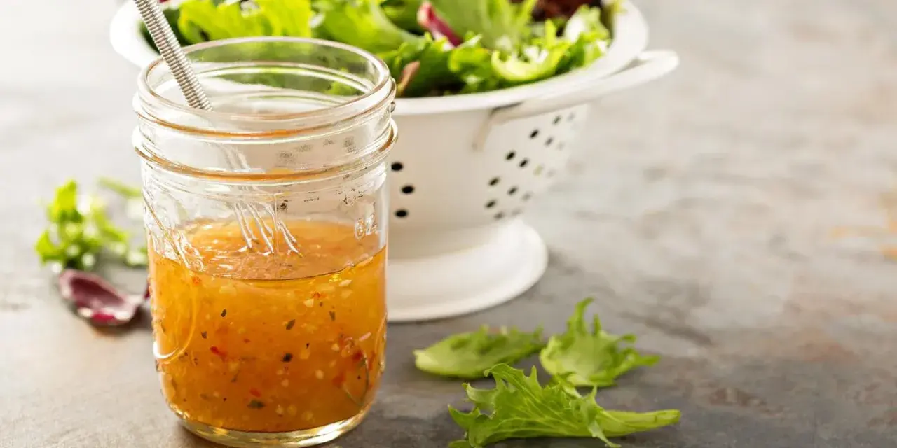 Ein Glas mit selbstgemachtem Salatdressing, das aussieht wie im Restaurant. Daneben ein Sieb mit frischem Salat.