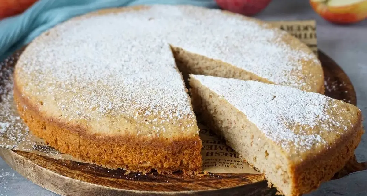 Ein schneller, saftiger Kuchen mit Apfelmus, bestäubt mit Puderzucker. Ein Stück wird gerade herausgenommen.