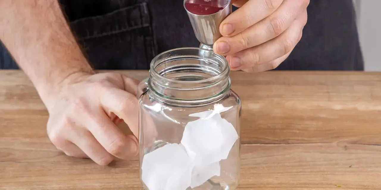 Eiswürfel in einem Glas, dazu wird rote Flüssigkeit aus einem Messbecher gegossen. So einfach lässt sich Rhabarberschorle selber machen!