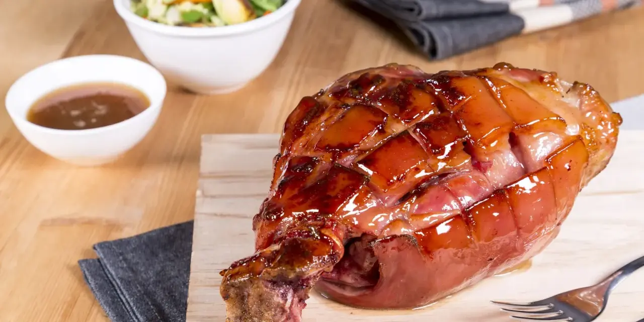 Glänzende, karamellisierte Schweinshaxe sous vide auf Holzbrett, daneben Schüssel mit Sauce und Salat.