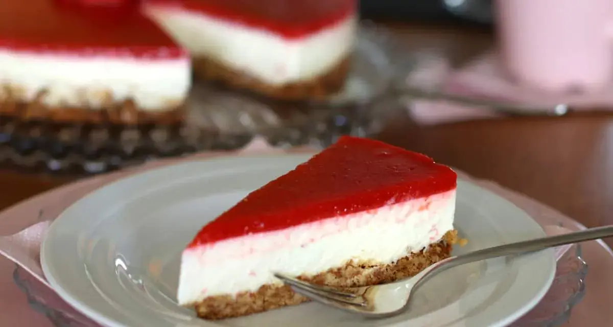 Ein Stück New York Cheesecake ohne Backen mit rotem Fruchttopping auf einem Teller, daneben ein ganzer Kuchen.