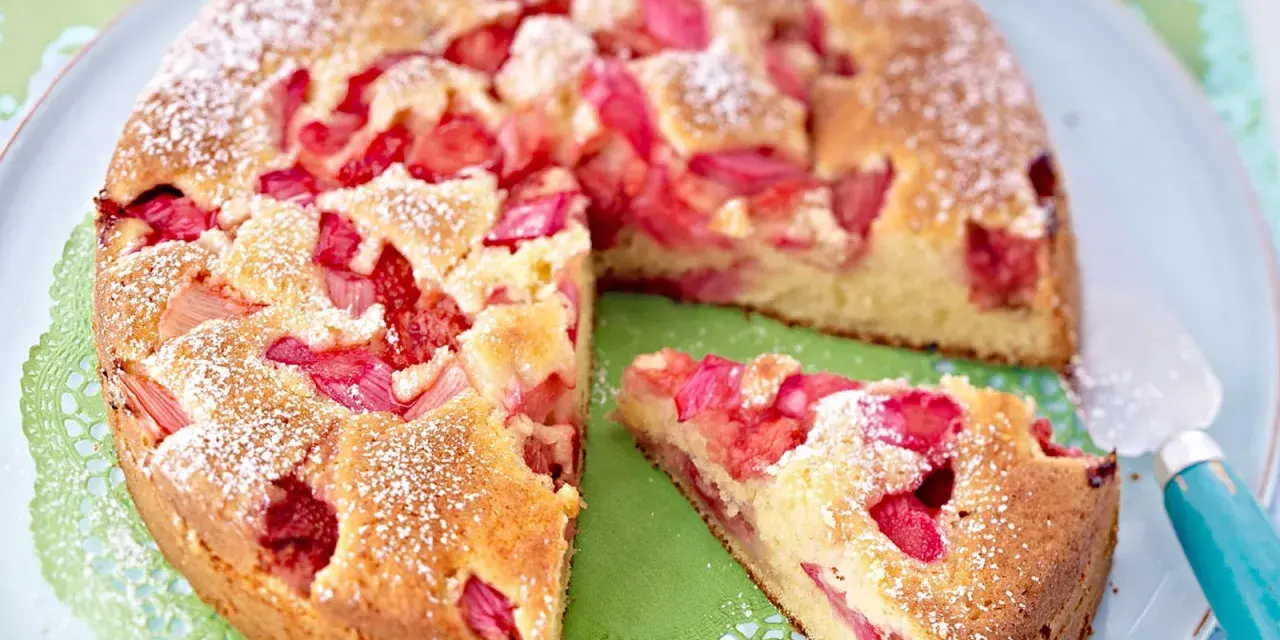 Ein saftiger Erdbeer Rhabarber Kuchen, bestäubt mit Puderzucker, wird auf einem Teller präsentiert. Ein Stück ist bereits herausgeschnitten.