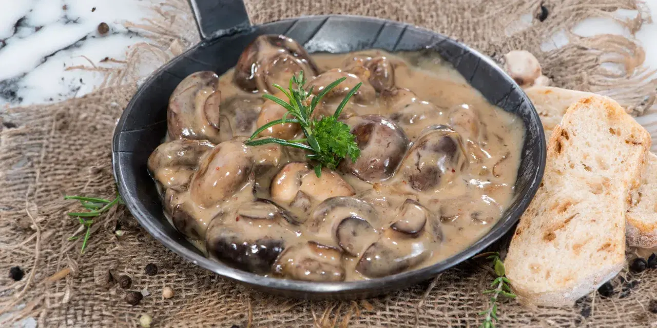 Herzhafte Champignon Rahmsauce in einer Pfanne, garniert mit Rosmarin, daneben knuspriges Brot und Pfefferkörner.