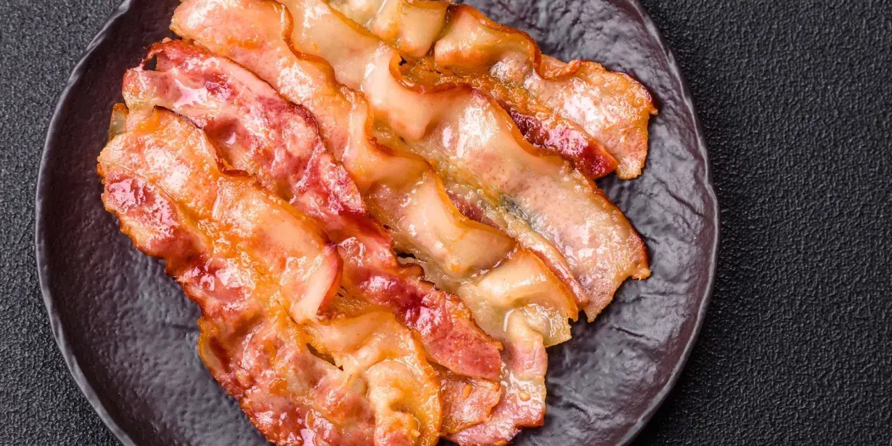 Knusprig gebratener Bacon im Ofen, auf einem dunklen Teller angerichtet.