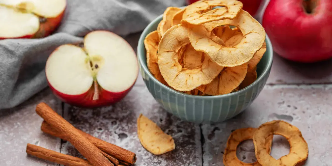 Leckere Apfelchips, die man ganz einfach selber machen kann, in einer Schüssel mit frischen Äpfeln und Zimtstangen.
