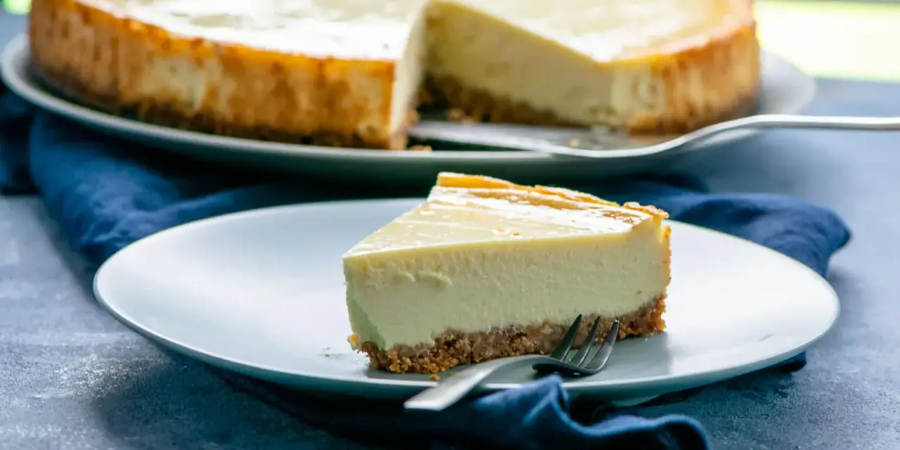 Ein Stück cremiger american cheesecake mit knusprigem Boden auf einem Teller, daneben ein ganzer Kuchen.