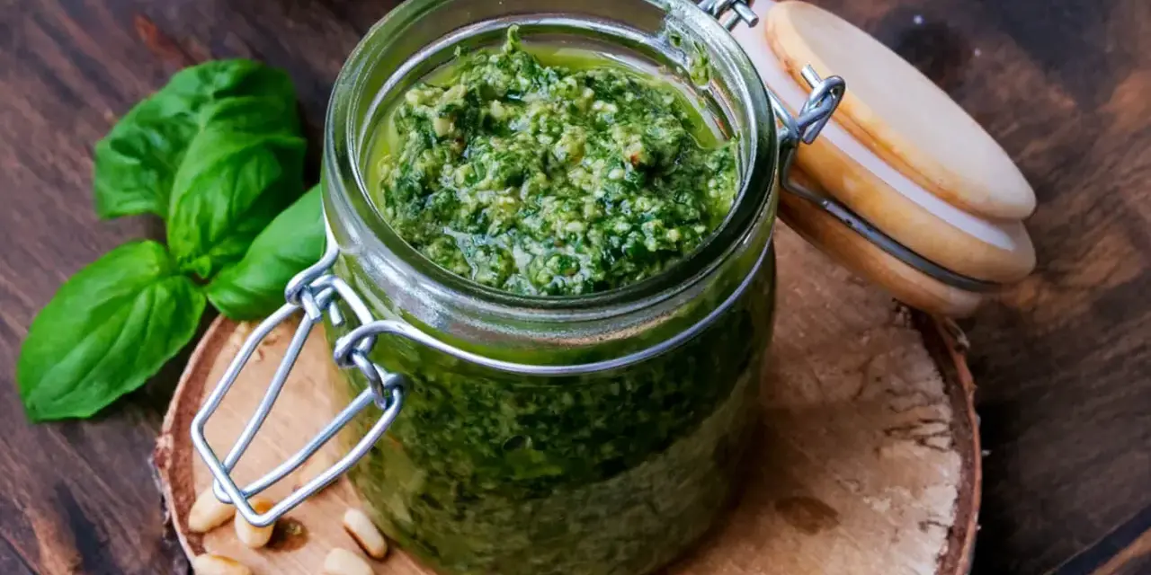 Frisches Basilikum Pesto in einem Glas, umgeben von Pinienkernen und Basilikumblättern.